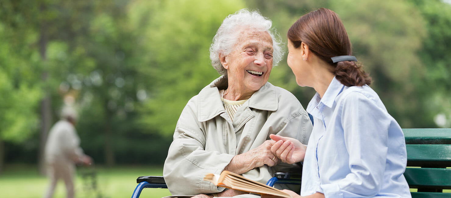 older woman sitting in wheelchair smiling and holding hands with younger woman sitting on bench with open book